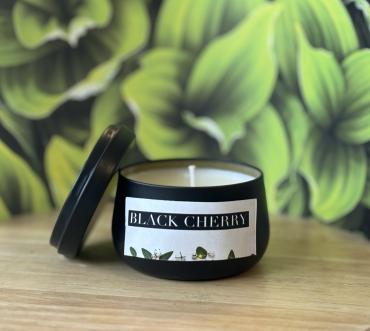 A black cherry candle in a black tin with a blurred foliage background