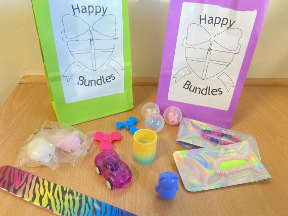 Various small toys are placed in front of two bags labelled 'happy bundles'. The toys are varied and consist of many different small trinkets, such as a mini slinkie, a wind up car, pencil toppers inside plastic capsules, mochi squishies in plapstic wrapping, a rainbow and black animal print slap-on bracelete and 2 small fidget spinners, a wobble toy, and finally, two hand-crafted bead bracelets.