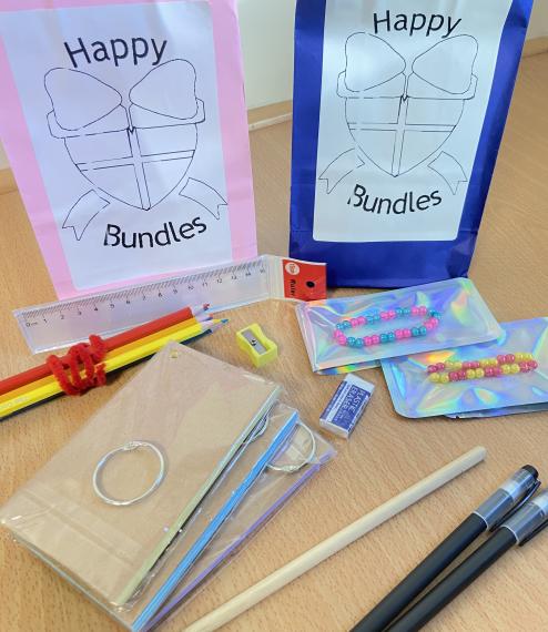 Various stationery are placed in front of two bags labelled 'happy bundles'. The stationery consists of a ruler, a pencil, writing pens, colouring pencils, a pencil sharpener and flashcards. Additionally there are two brightly coloured bead bracelets.