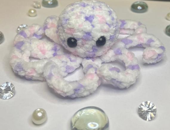 The image shows a small, handmade plush octopus sitting on a light surface. It appears to be crocheted or knitted from very soft, fluffy yarn. The octopus is mostly white with pastel purple and pink patches blended throughout, giving it a gentle, marbled look.  It has large, shiny black eyes that give it a cute and expressive appearance. Its eight tentacles are short, rounded, and slightly curled, arranged around its round body.