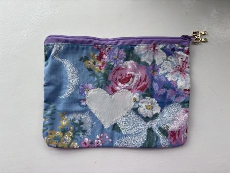 pouch using blue fabrics with different coloured flowers on white heart sewn onto front, purple zip.