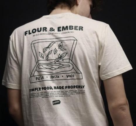 A person seen from behind wearing a relaxed-fit, cream-coloured t-shirt against a dark background. The shirt features a bold, hand-drawn graphic print across the full back in dark green ink, including large display typography, a line-art illustration, and multiple text blocks of varying sizes. A small rectangular brand logo reading "Palette" is printed at the centre bottom of the design, serving as the maker's mark.